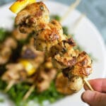 One skewer with grilled chicken and lemons held close to the camera with plate in the background.